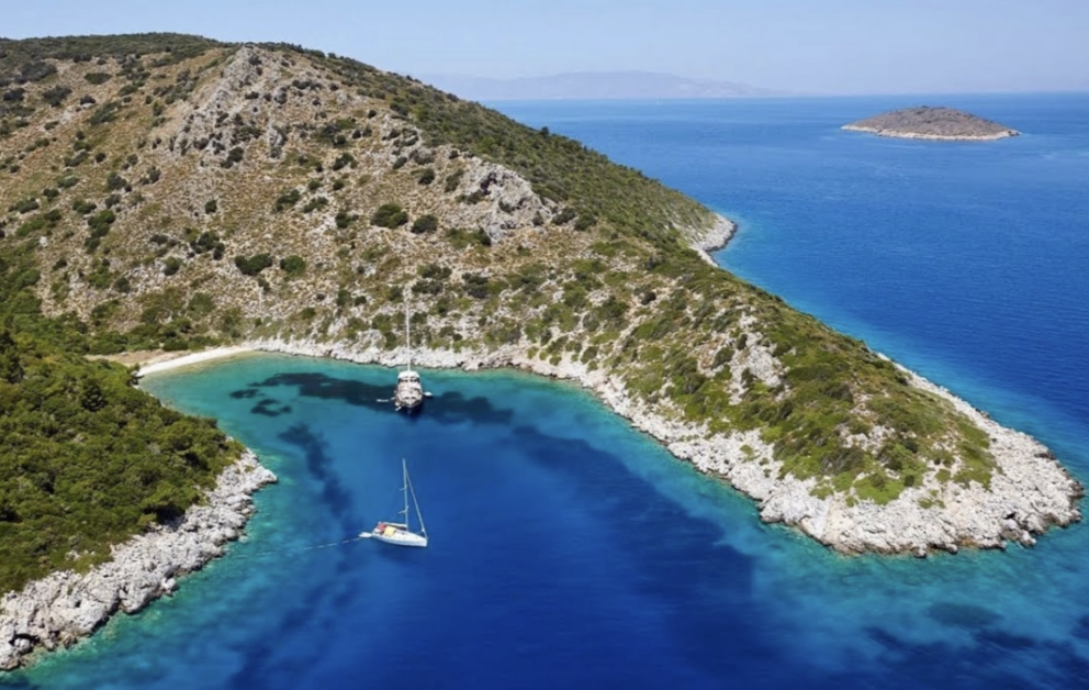 A high-angle shot of a rocky, green peninsula extending into a turquoise sea. Two boats, a large motor yacht and a smaller sailboat, are anchored in a sheltered cove with a small sandy beach. A small, uninhabited island is visible in the distance under a clear blue sky.