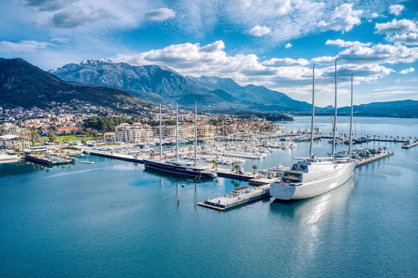A large marina in Porto Montenegro, with several yachts docked. Two very large, modern sailing yachts are prominent in the foreground, with one of them, the Sailing Yacht A, recognizable by its unique design. The marina is surrounded by a coastal town at the base of large, snow-capped mountains under a partly cloudy sky. The water in the bay is calm and blue.