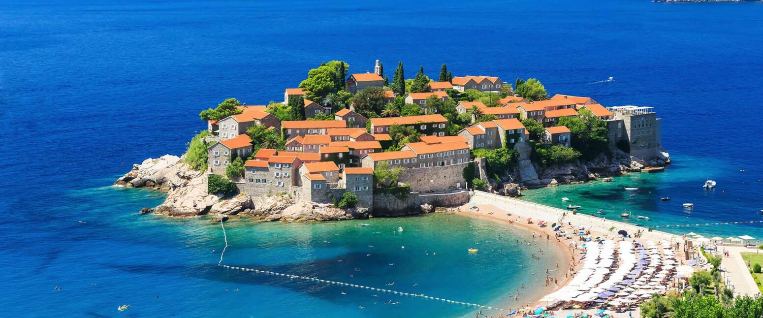 A panoramic view of Sveti Stefan, a small islet in Montenegro, connected to the mainland by a narrow causeway. The island is covered with old stone buildings with red roofs, surrounded by the deep blue Adriatic Sea. A beach with sun loungers and umbrellas is visible on the right side of the causeway.
