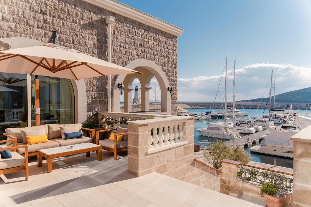 A stone terrace with a beige sofa and chairs under a large umbrella, overlooking a marina with boats and yachts. In the background, there is a stone building with arched openings and a view of the sea and mountains.
