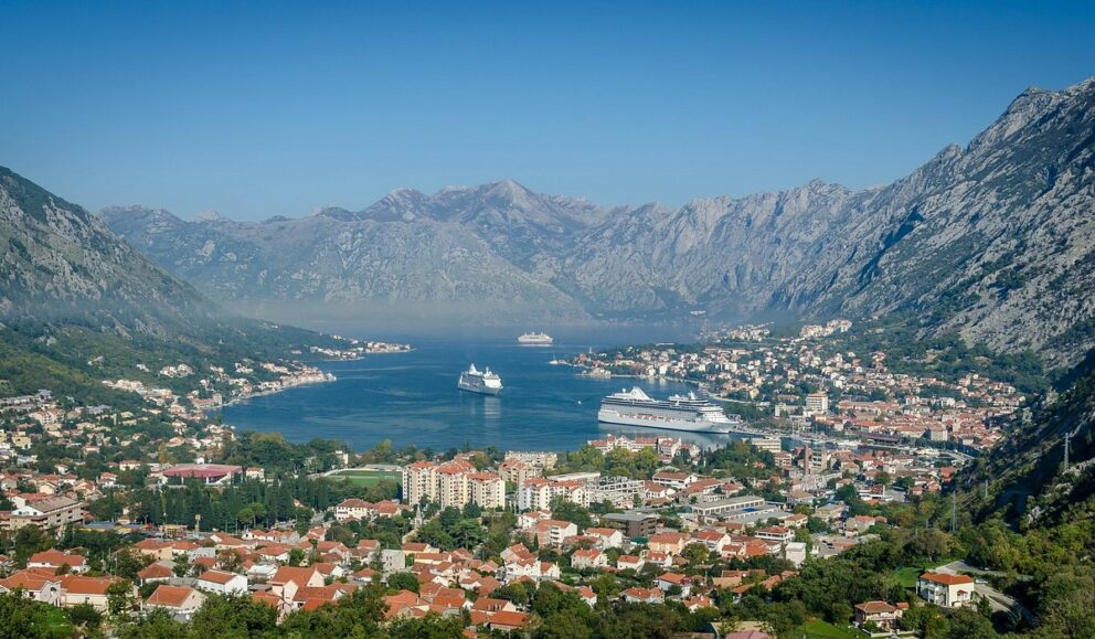 Scenic view of the Bay of Kotor in Montenegro. The bay is framed by large, imposing mountains and has a town with numerous buildings, many featuring red roofs, situated along the shoreline. Several large cruise ships are visible in the clear blue water of the bay. The sky is a clear blue, suggesting a sunny day.