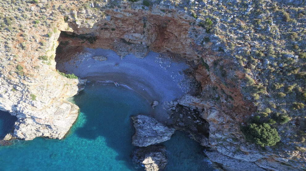 Aerial view of a small, secluded cove with a grey pebble beach known as Red Rock Beach in Montenegro. The cove is surrounded by steep, rocky cliffs, and the clear turquoise water leads into a large cave opening on the left side.