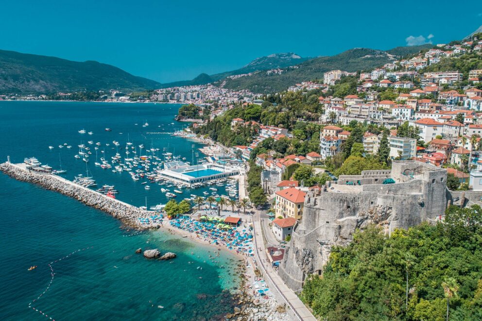 A marina filled with numerous boats docked behind a long stone breakwater. A small beach with sun loungers and umbrellas. The Forte Mare, a historic fortress, situated on a rocky outcrop overlooking the sea. A dense collection of red-roofed buildings and houses stretching up the steep hillside. Lush green mountains in the background under a clear blue sky.