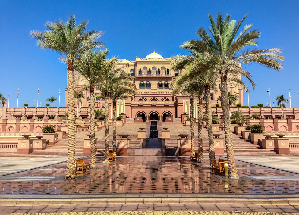 The grand Emirates Palace in Abu Dhabi stands under a clear blue sky, framed by tall palm trees and elegant stairways leading to its arched entrance. The polished courtyard reflects the sunlight, emphasizing the building’s golden-brown tones and intricate architecture.