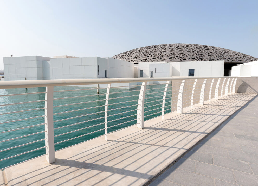 Modern architectural view of the Louvre Abu Dhabi with its distinctive geometric dome partially covering white cube-like buildings, surrounded by calm turquoise water under a clear blue sky.