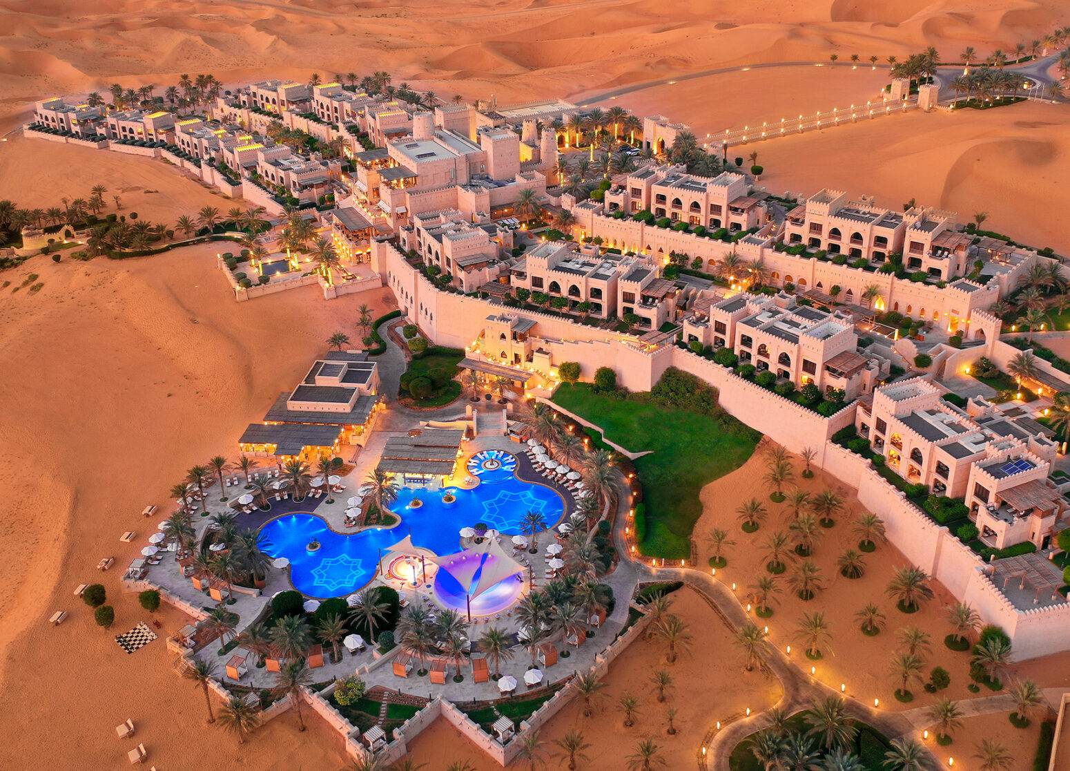 Aerial view of the luxurious Qasr Al Sarab Desert Resort in Abu Dhabi, featuring an oasis-like pool surrounded by palm trees, lush gardens, and sand-colored buildings blending into the vast golden desert landscape at sunset.