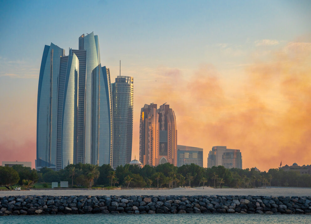 View of the Abu Dhabi skyline at sunset, featuring modern high-rise towers with reflective glass facades, palm trees along the shoreline, and soft orange clouds in the background above the calm Arabian Gulf waters.