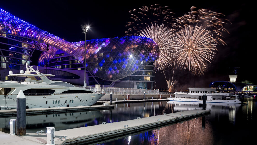 Fireworks light up the night sky over a modern marina in Abu Dhabi, reflecting on calm waters. Luxury yachts are docked in front of a futuristic, illuminated building with a curved, glowing blue and purple facade.