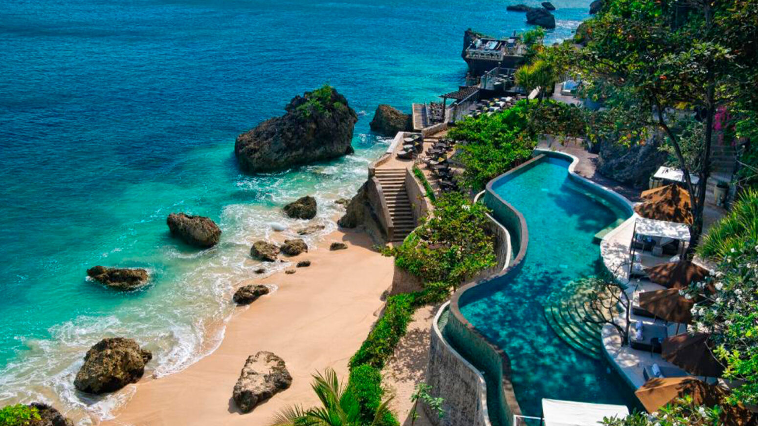 Luxury cliffside resort in Bali with an infinity pool overlooking a turquoise ocean and a sandy beach dotted with rocks and tropical greenery.