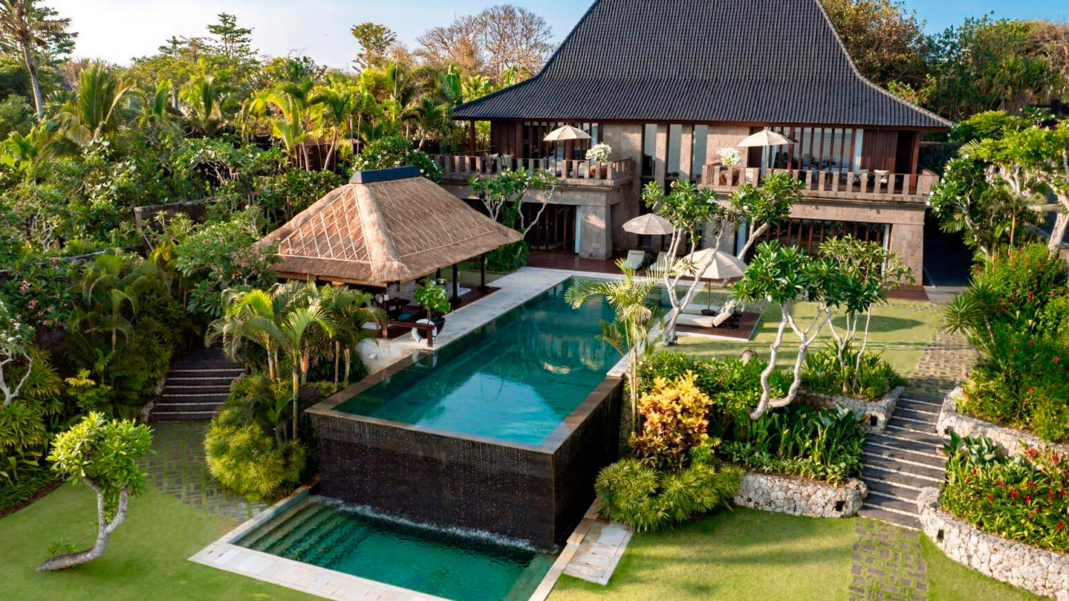 Luxurious Balinese villa surrounded by lush tropical gardens, featuring a private infinity pool, traditional thatched gazebo, and elegant two-story architecture with wooden accents.
