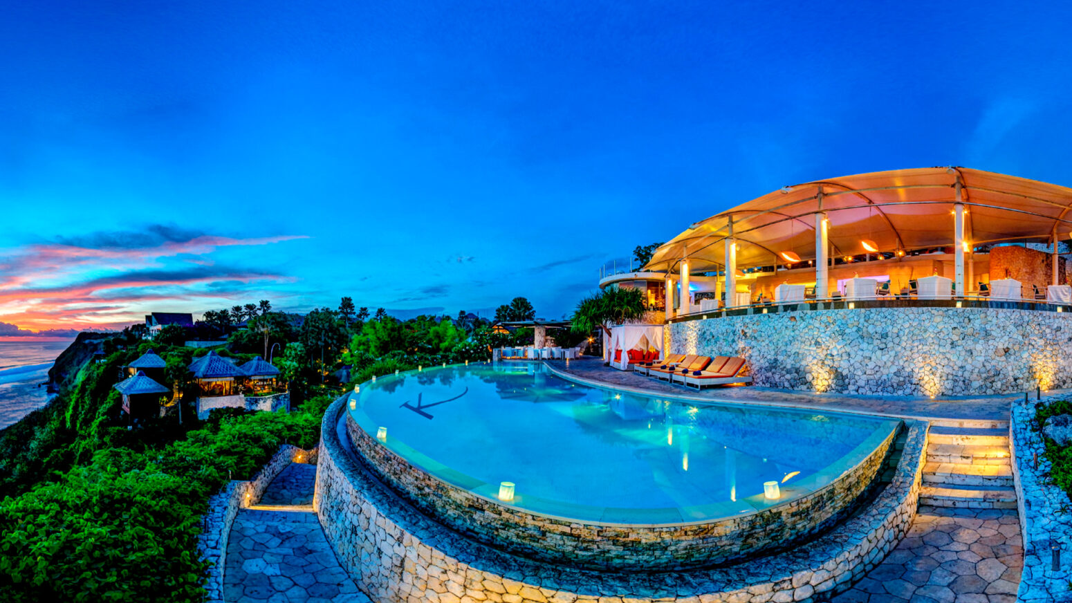 Elegant cliffside resort in Bali with a curved infinity pool overlooking the ocean at sunset, surrounded by stone terraces, cabanas, and soft evening lighting.