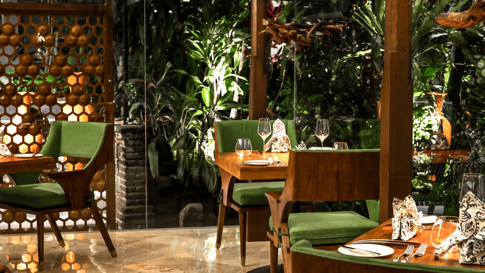 Elegant restaurant interior in Bali featuring wooden tables, green cushioned chairs, soft lighting, and lush tropical plants visible through glass walls, creating a serene dining atmosphere.