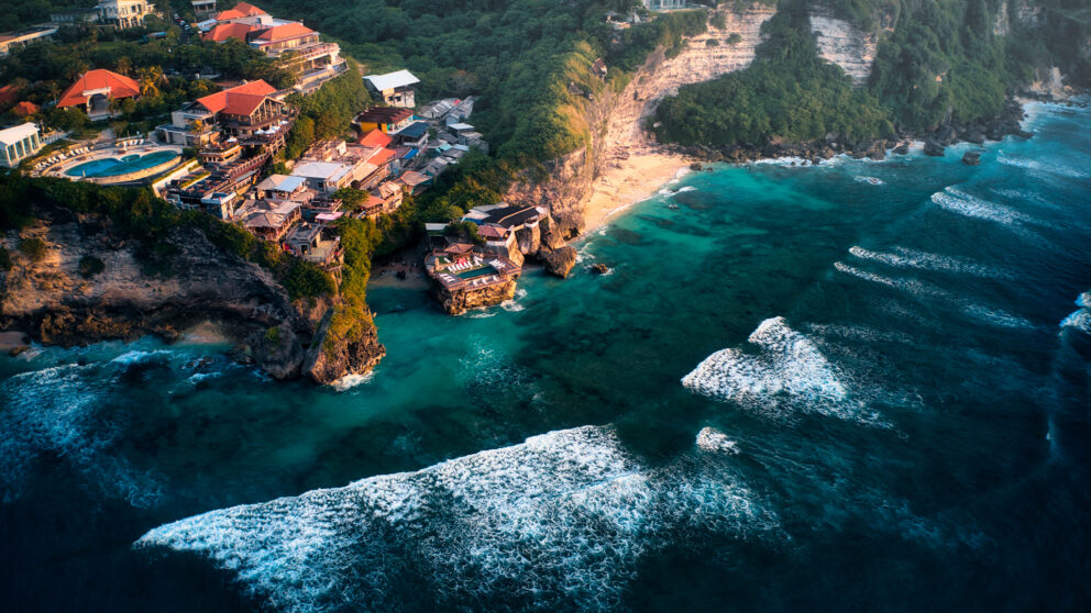 Aerial view of Uluwatu, Bali, showing turquoise ocean waves crashing against dramatic cliffs lined with villas and beachside bars overlooking a hidden sandy cove.