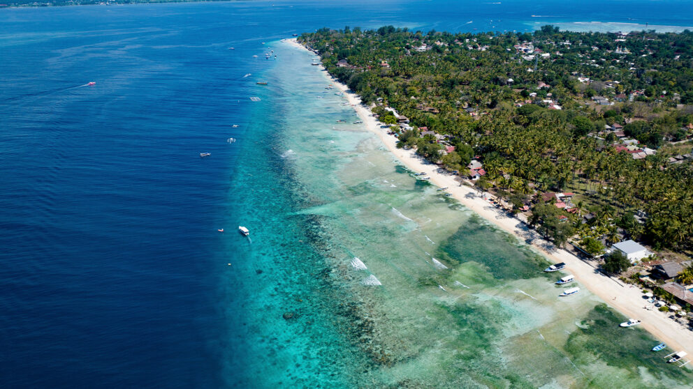 Aerial view of Gili Island, Indonesia, showing turquoise shallows meeting deep blue ocean waters, white sandy beaches, small boats, and lush greenery dotted with coastal villas.