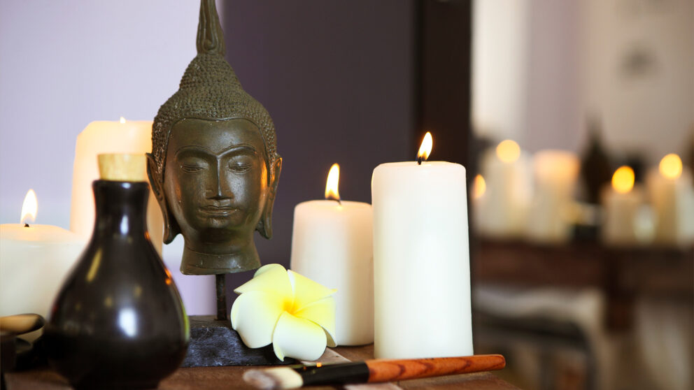 Close-up of a peaceful spa scene with a Buddha statue, burning candles, a frangipani flower, and essential oil bottles, creating a serene and relaxing atmosphere.
