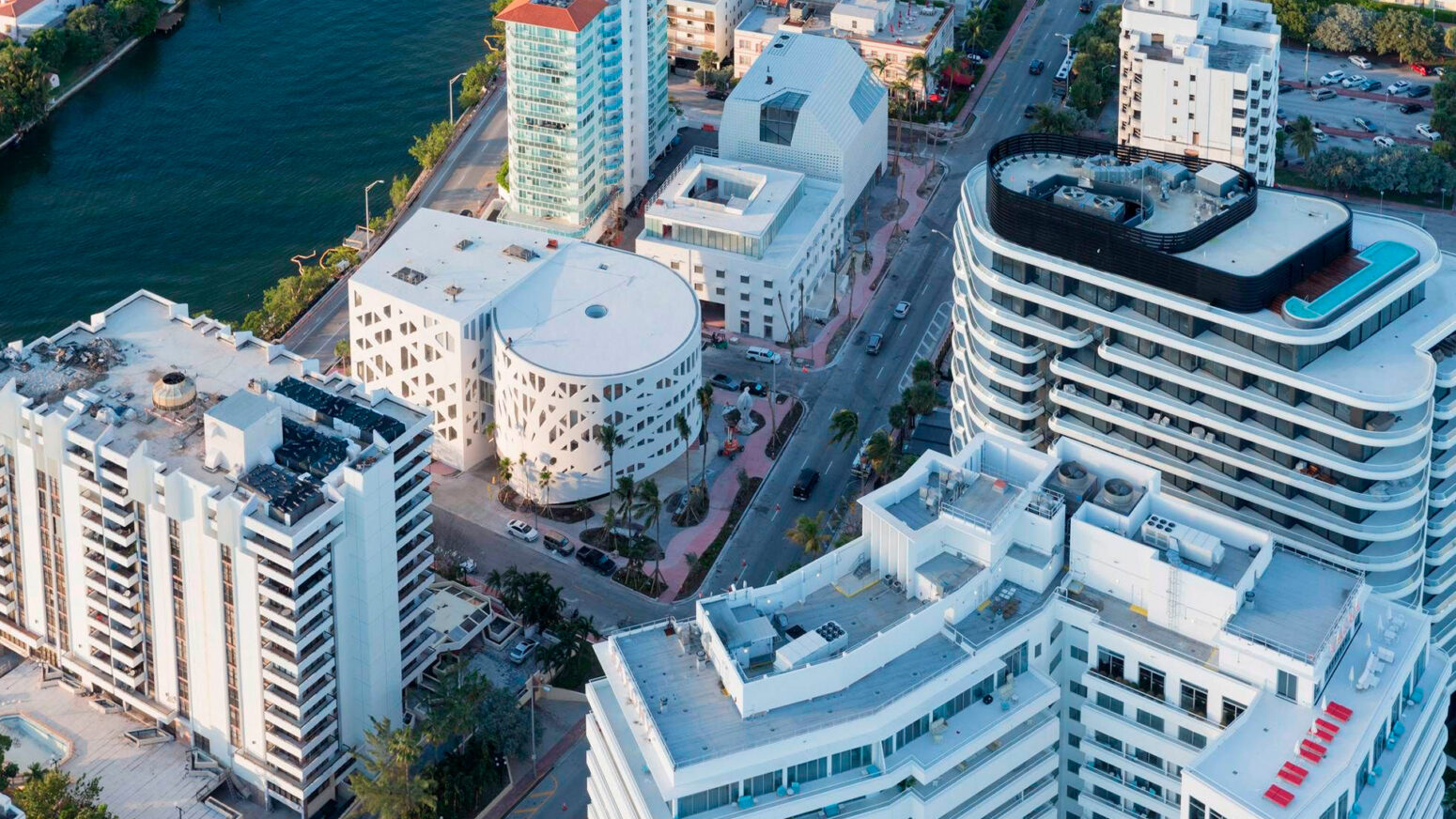 An aerial view of several modern, white buildings in a city with a body of water nearby. The buildings have unique architectural designs, including a circular building with a patterned facade and others with terraced roofs. Palm trees and a street with cars are visible below. The scene depicts the Faena District in Miami Beach.