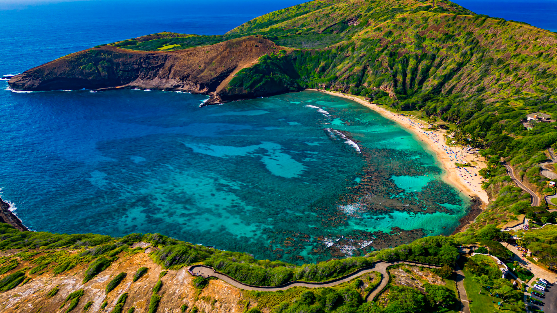 A scenic aerial view of Hanauma Bay, a crescent-shaped bay on the island of Oahu, Hawaii. The image shows the bay's clear turquoise water with visible coral reefs, a sandy beach with people, and the surrounding green and brown volcanic crater rim. A winding road is visible on the hillside leading down to the bay.