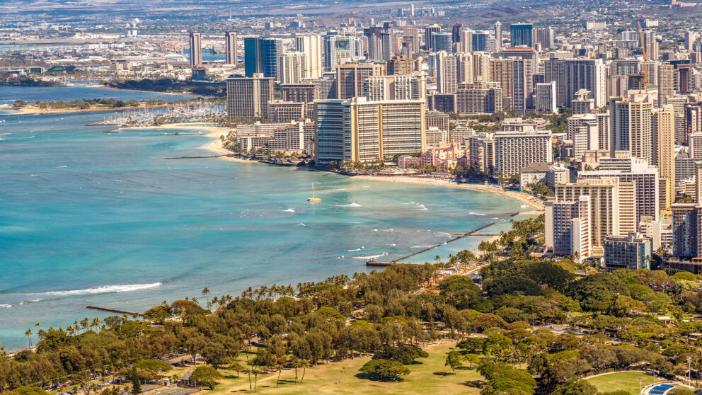 A high-angle shot of Waikiki Beach in Honolulu, Hawaii, showing the coastline with turquoise water, a sandy beach, and a dense urban skyline of high-rise buildings in the background. In the foreground, there is a green park with palm trees.