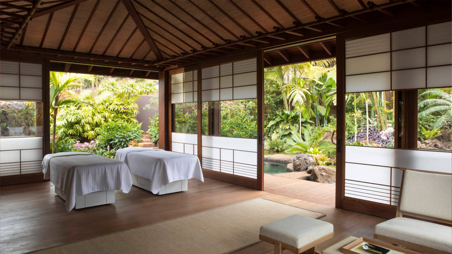 A tranquil spa room with two massage tables, a wooden floor and ceiling, and large sliding doors that open to a lush, tropical garden with a small pond.