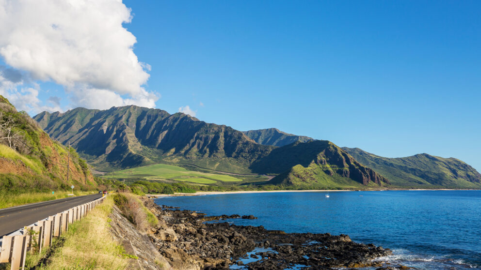 A coastal road winds along a rocky shoreline next to a calm blue ocean. In the background, large, lush green mountains rise under a clear blue sky with some white clouds.