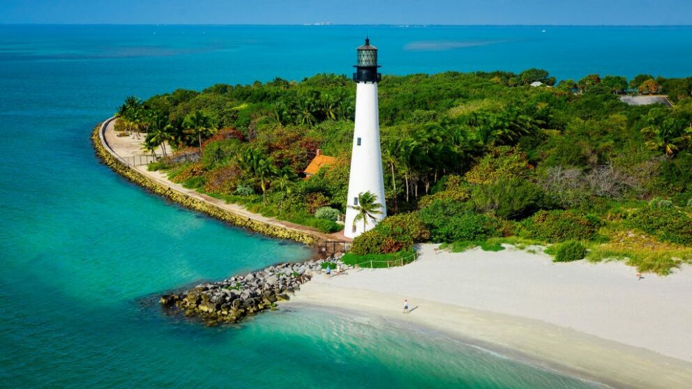 A white lighthouse with a black lantern room stands on a sandy peninsula surrounded by turquoise water and lush green trees. The image, taken from an aerial perspective, shows the lighthouse at Bill Baggs Cape Florida State Park.