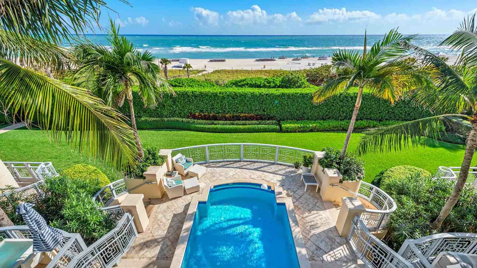 A high-angle view of a luxury property's backyard, featuring a swimming pool and patio in the foreground, leading to a manicured green lawn with palm trees. In the background, a sandy beach and the ocean are visible under a partly cloudy sky.