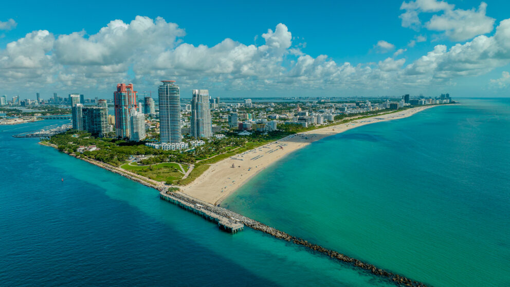An aerial view of South Pointe Park in Miami Beach, Florida, showing a sandy beach, a long rock jetty, and several high-rise buildings along the coastline. The water is a clear turquoise and the sky is blue with scattered clouds.