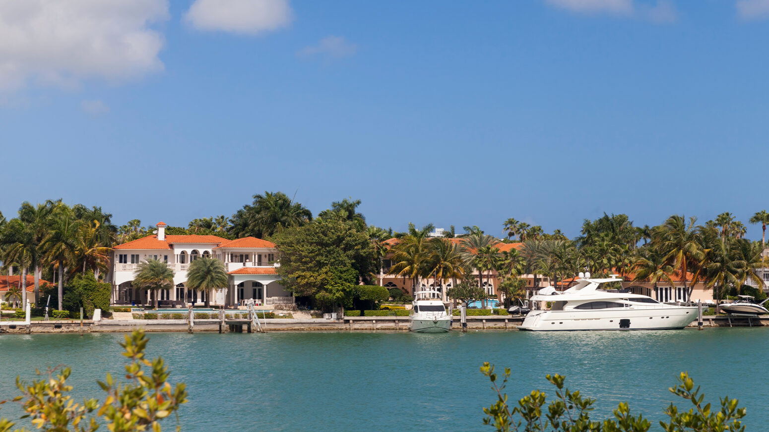 A bright, daytime image shows a large, luxurious waterfront mansion in a tropical setting. The house is a multi-story, white building with a red-tile roof and large windows, surrounded by palm trees and lush vegetation. A substantial white yacht is moored at a private dock in the foreground