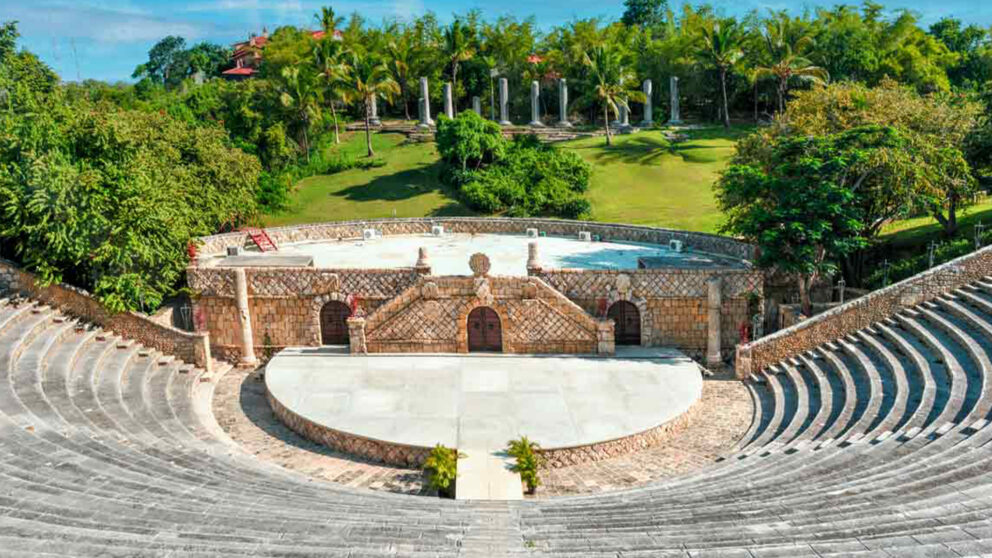 Stone amphitheater surrounded by lush green trees and palm trees under a clear blue sky.