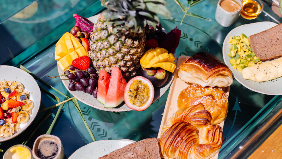 Colorful brunch table with tropical fruits, pastries, bread, and coffee served on a glass table in natural light.