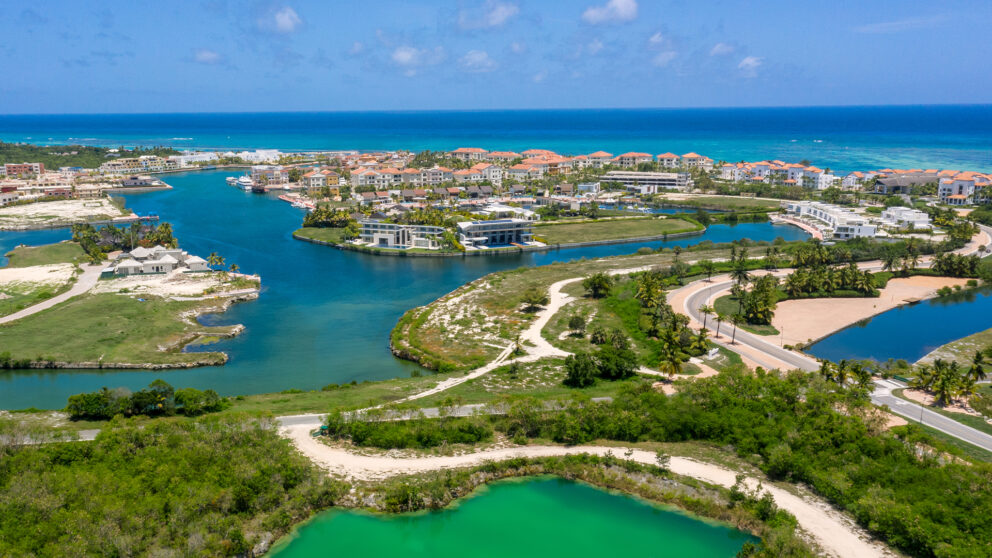 Aerial view of a luxury coastal resort with canals, villas, and green landscapes near the turquoise ocean under a bright blue sky.