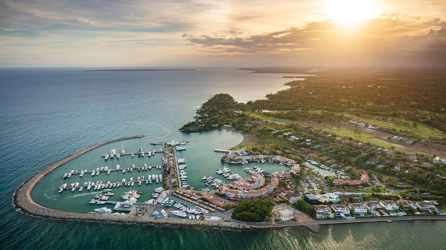 Aerial view of a marina with yachts, waterfront buildings, and lush green landscapes at sunset by the ocean.