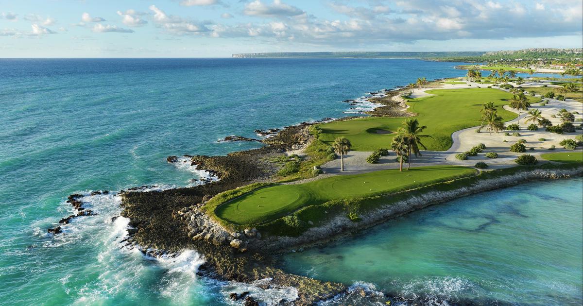 Aerial view of a scenic oceanfront golf course with green fairways, palm trees, and turquoise water along a rocky coastline.