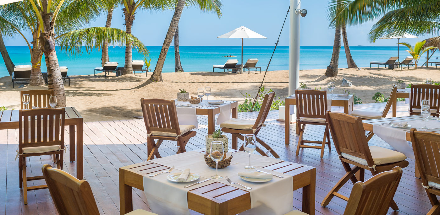 Beachfront restaurant with wooden tables, white tablecloths, and palm trees overlooking the turquoise ocean on a sunny day.