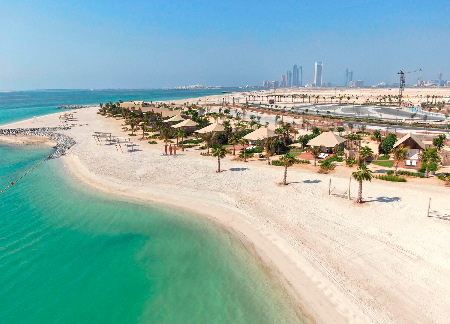 Aerial view of Al Hudayriyat Island in Abu Dhabi, featuring white sandy beaches, turquoise waters, rows of palm trees, and beachfront cabanas. The Abu Dhabi skyline is visible in the distance under a clear blue sky.