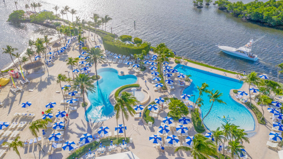 An aerial view of a luxurious resort with multiple swimming pools and a sandy beach area. The pools are surrounded by rows of lounge chairs with blue and white striped umbrellas. Palm trees are scattered throughout the property, which is situated next to a body of water with a boat docked nearby.